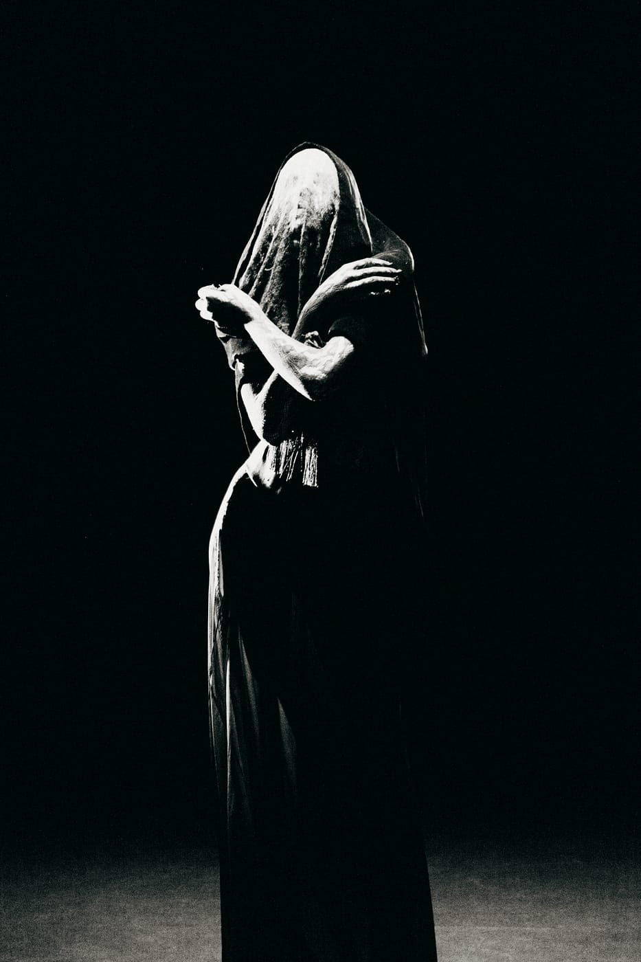 A veiled figure in a long dark gown, one arm raised across the face