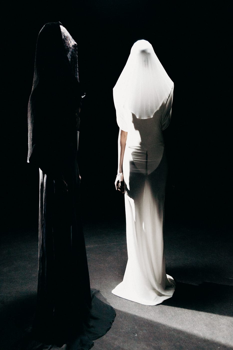 Two veiled figures, one in black and one in white, standing in a single shaft of light