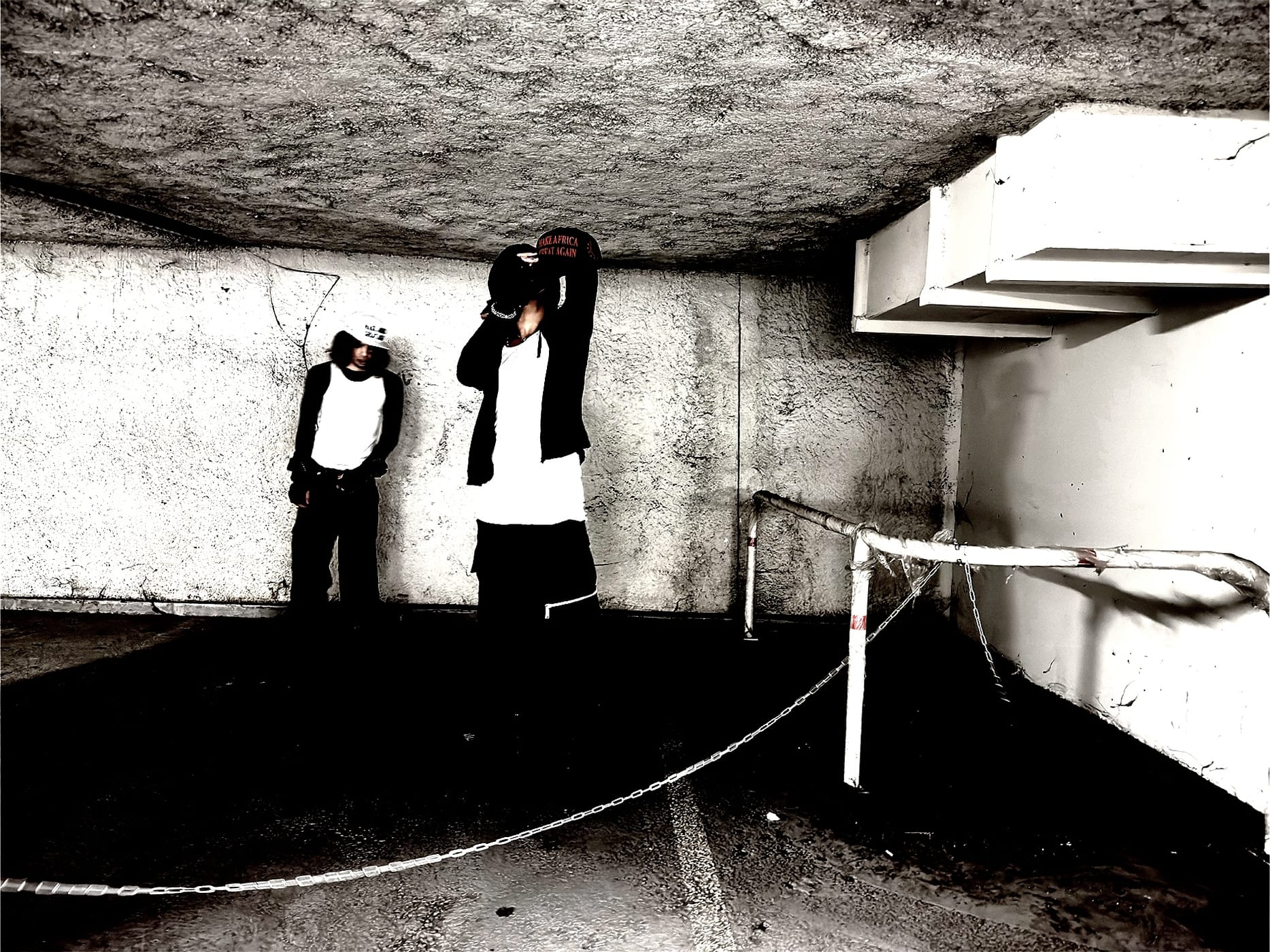 Two figures against a raw concrete wall in a basement, caps low
