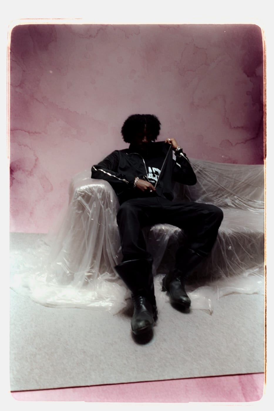 Peso Shabba seated on plastic sheeting against a wet pink ground, all in black