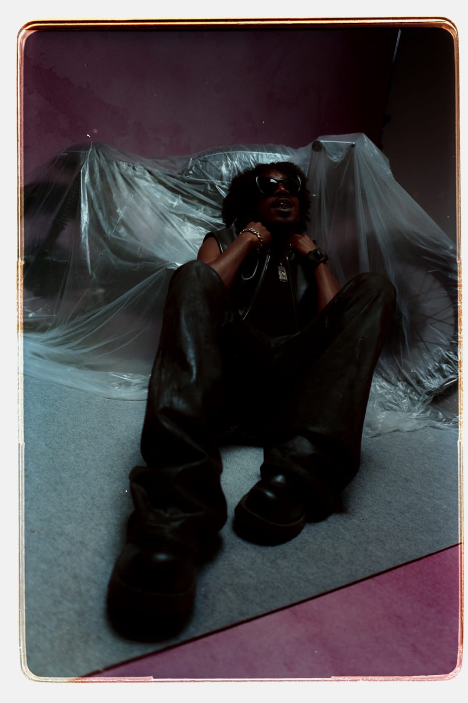 Peso Shabba on the floor in leather and shades, sheeting drawn up behind him