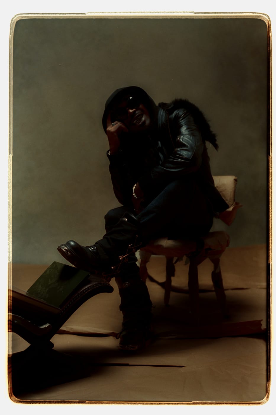 Peso Shabba seated in a fur-collared leather jacket, boot up on a broken chair