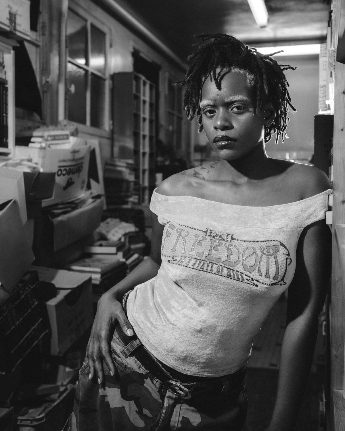 Ezek leaning against a shelf, short blonde-tipped locs, bleached brows, a velvet top reading Freedom