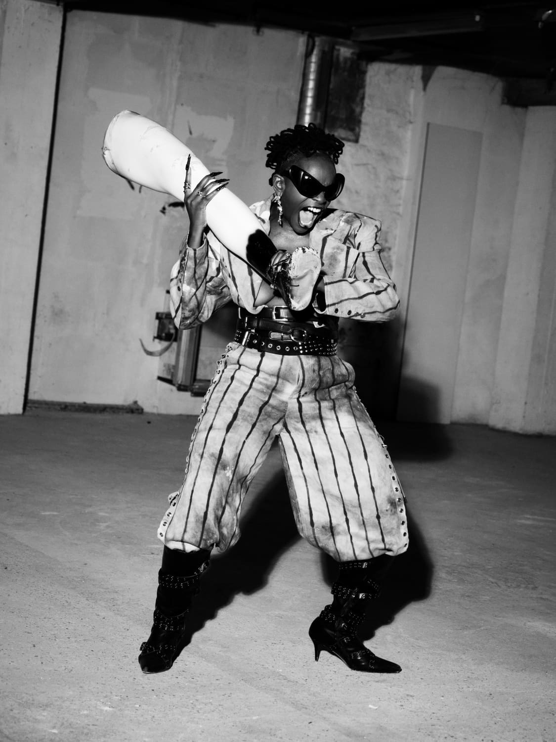 Ezek laughing in a striped suit and black corset belt, sunglasses on, a length of white pipe over her shoulder