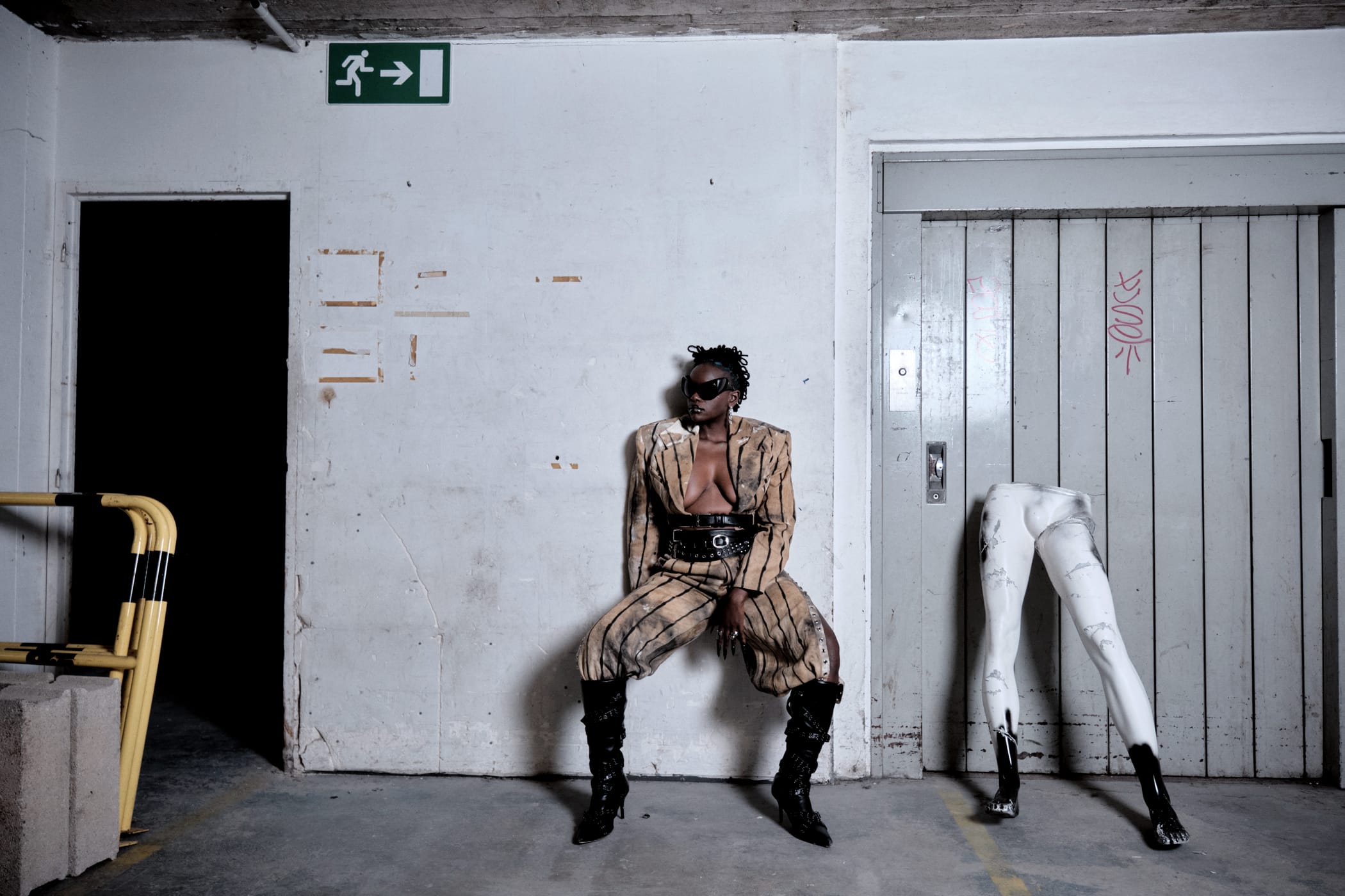 Ezek seated against the wall in a striped suit and black corset belt, sunglasses on, a pair of mannequin legs by the lift