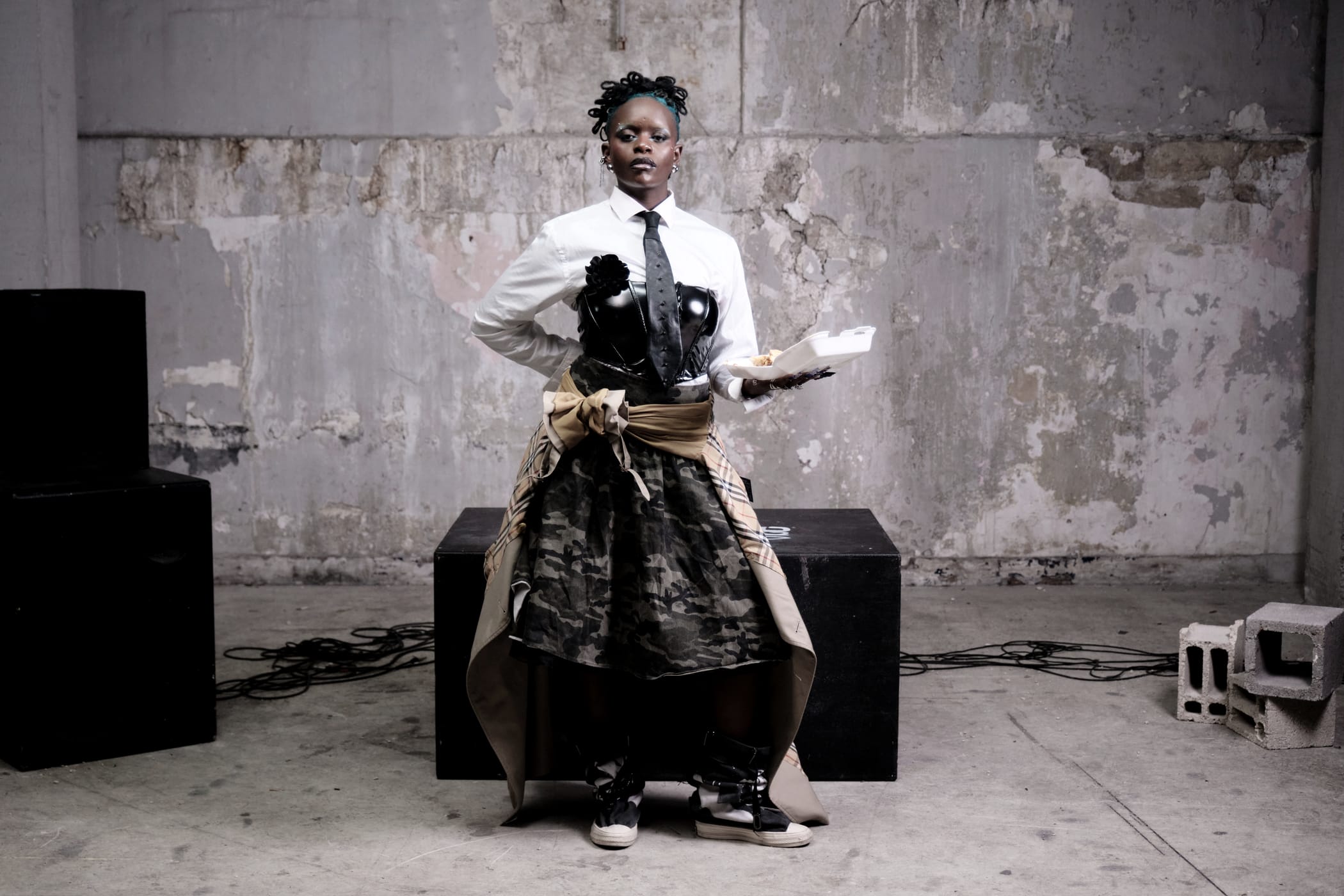 Ezek standing in white shirt, tie and camouflage skirt, arms open, in front of the black block