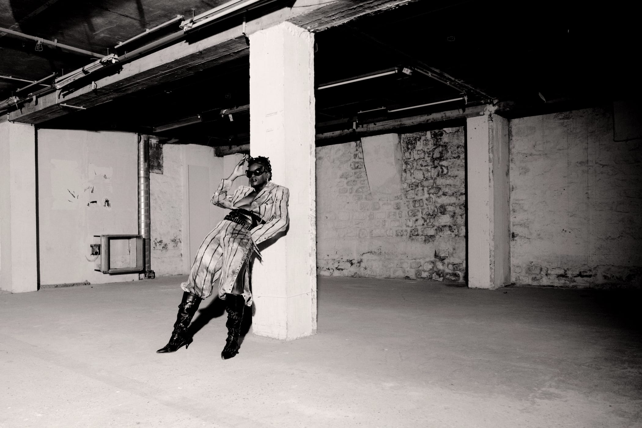 Ezek leaning back against a pillar in the striped suit, one hand to her brow, toned monochrome