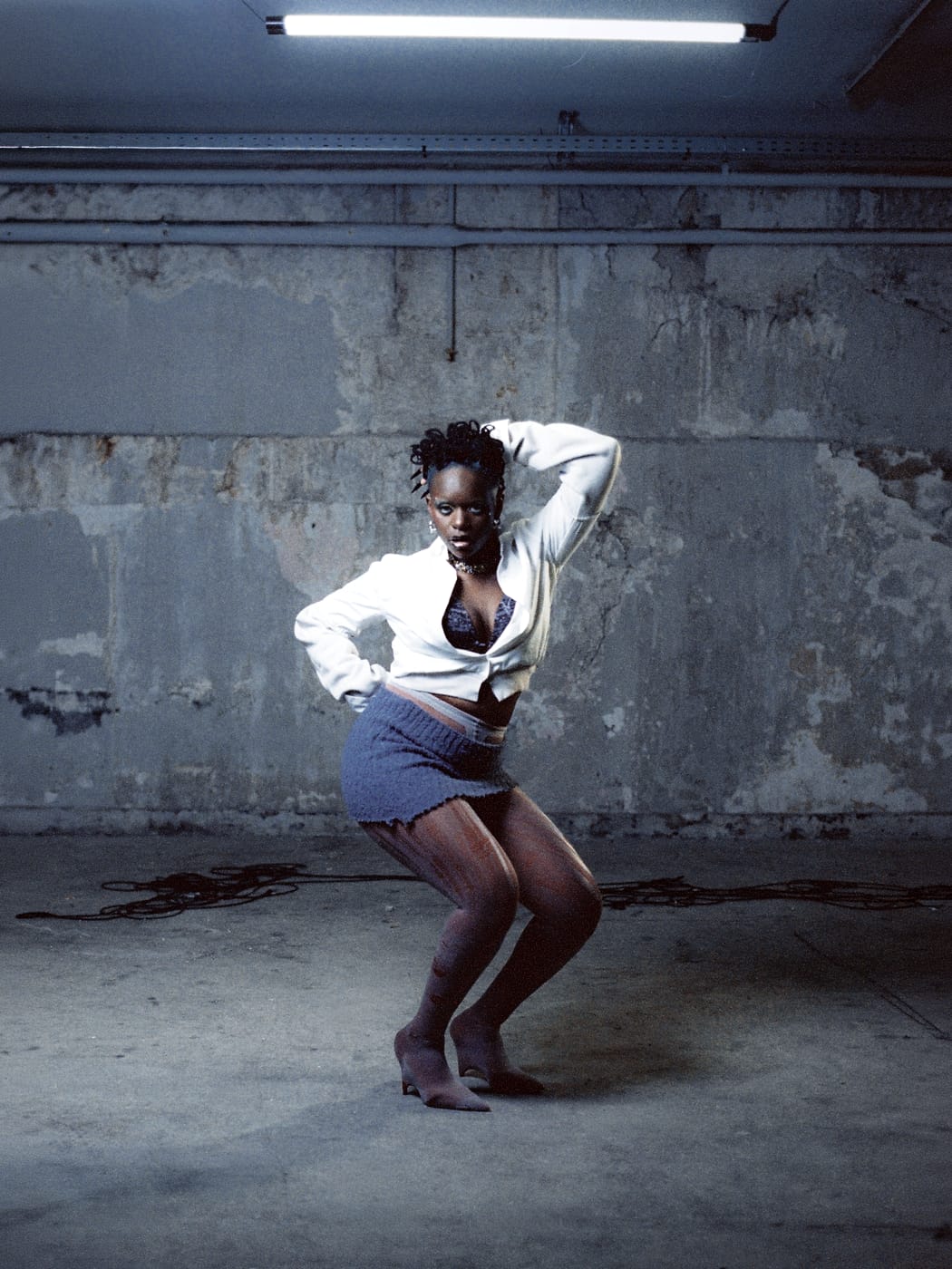 Ezek mid-dance under the strip light, white shirt open, knit skirt