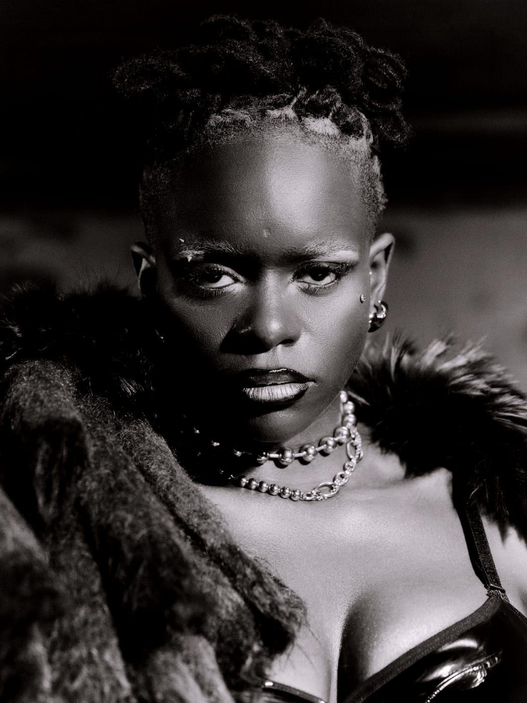 Ezek close, chains at her neck, fur at her shoulders, toned monochrome