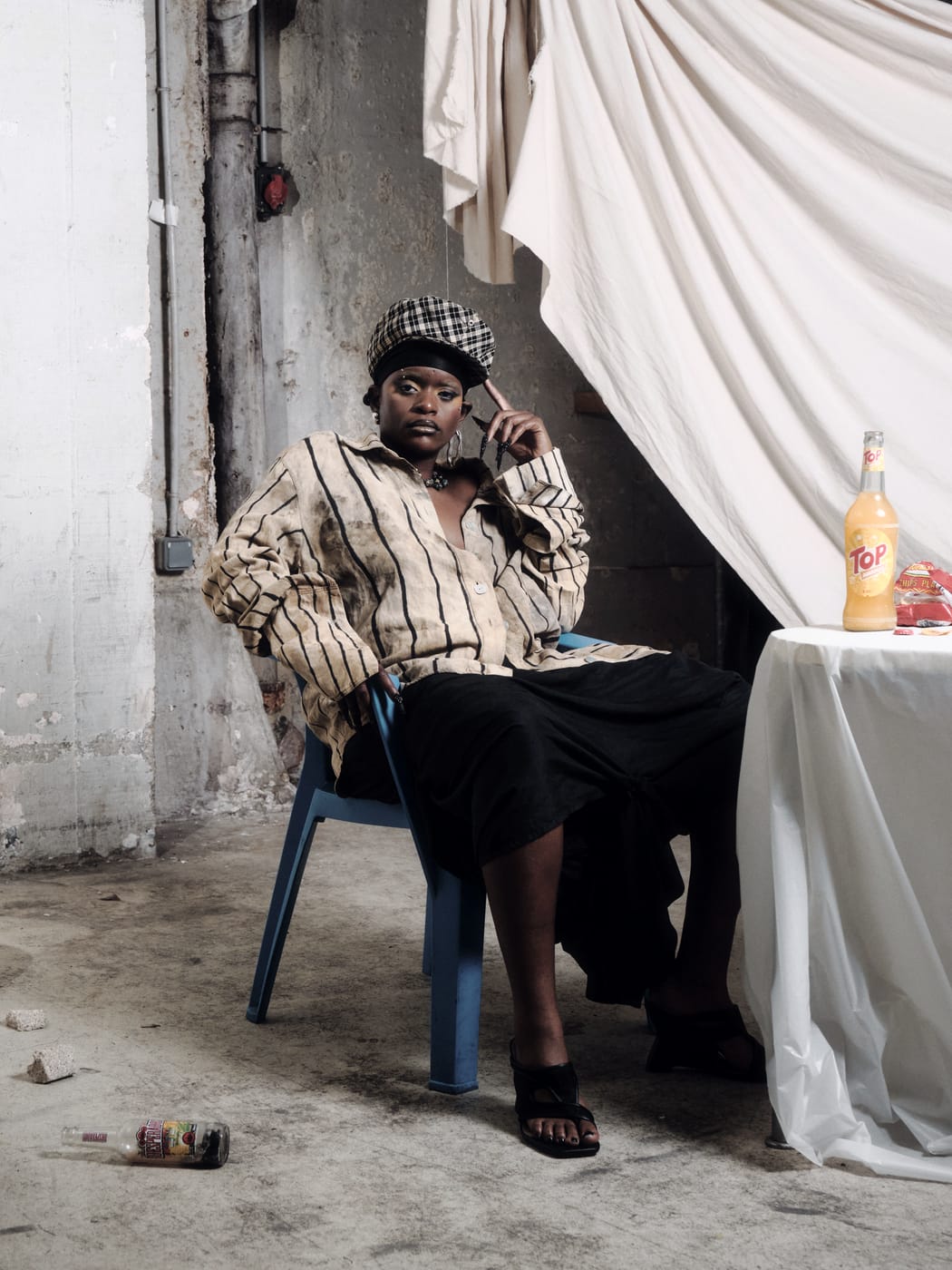 Ezek seated on the blue chair by the table, a bottle held to her ear, beret on