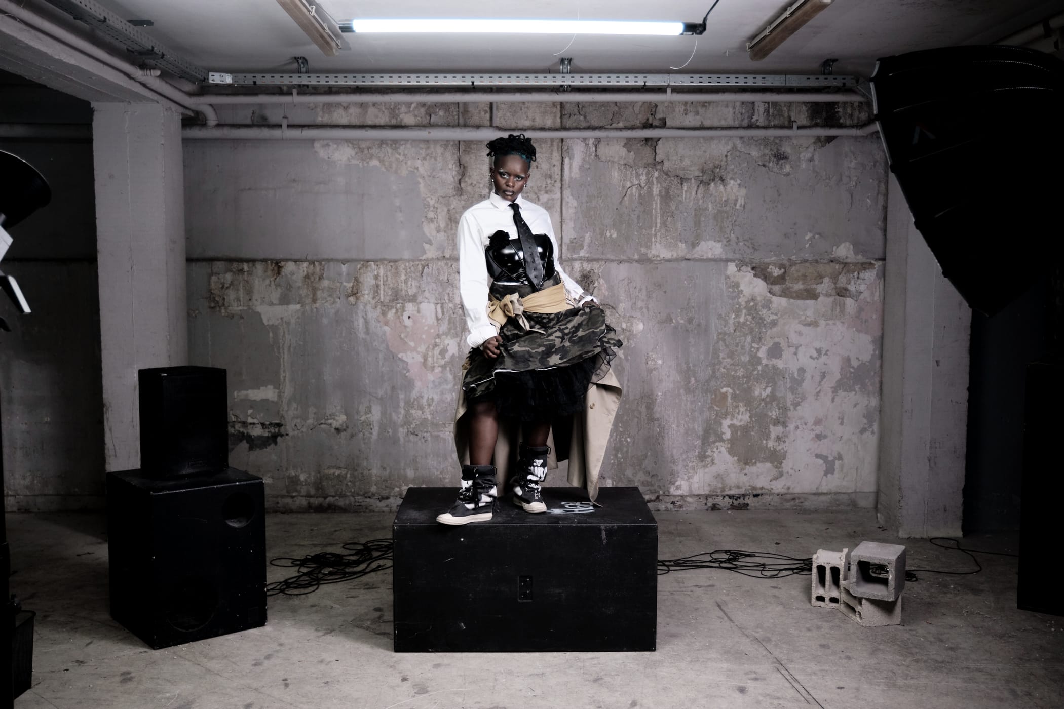 Ezek standing on the black box in shirt, tie and camouflage skirt, tulle at the hem