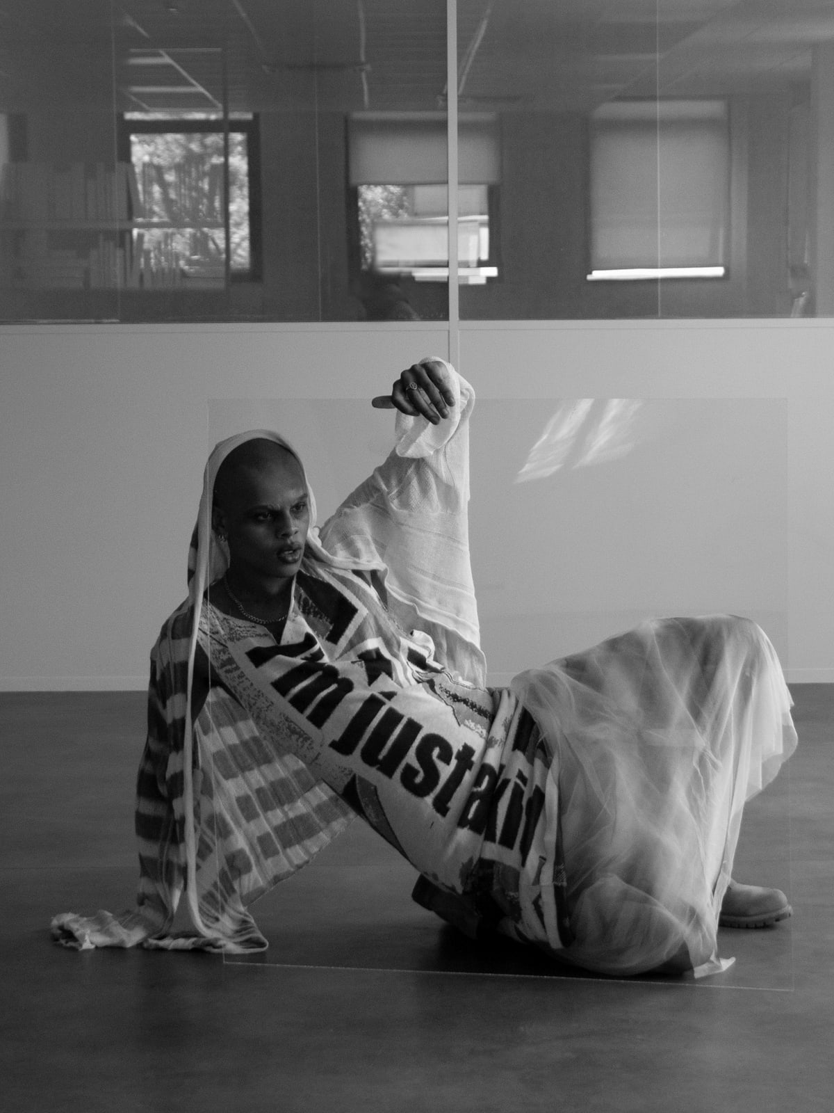 Oluwa seated on the floor behind a sheet of glass, hooded, one hand raised over its edge