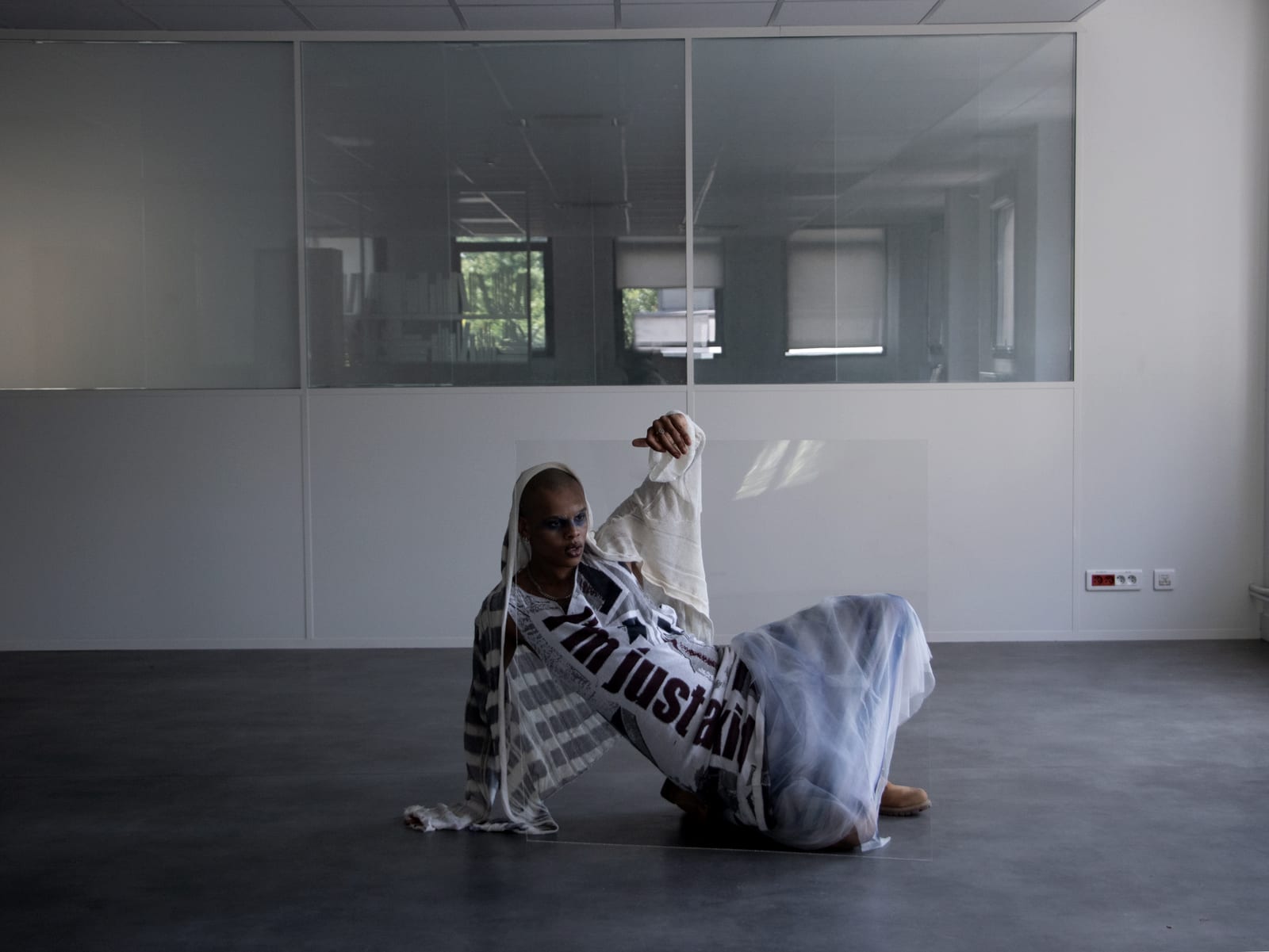 Oluwa seated on the floor behind a sheet of glass, one finger resting over its edge, in a tulle skirt and striped shawl