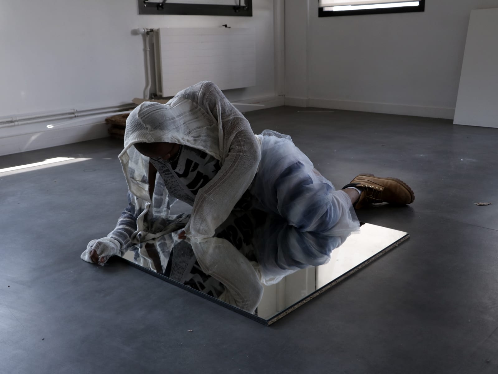 Oluwa crouched over a mirror laid on the floor, hooded