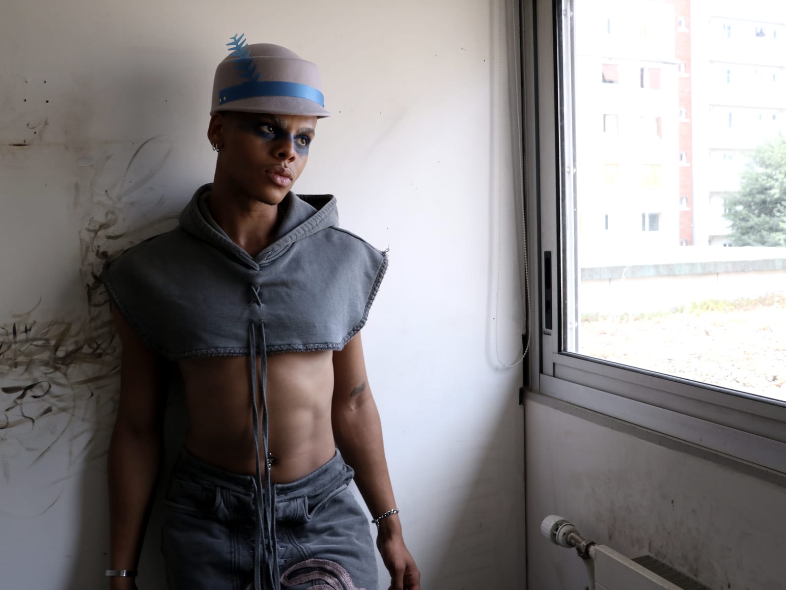 Oluwa standing in the cropped grey hood against a raw wall